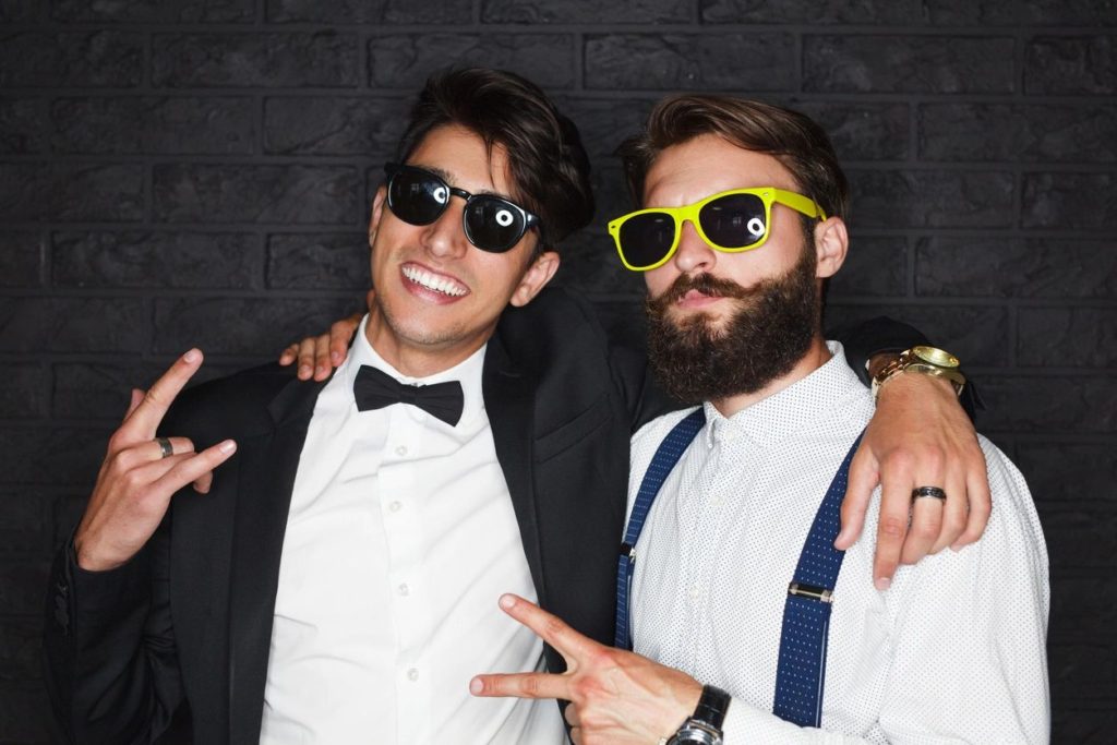 A couple of entitled looking young guys wearing tuxes and sunglasses making rap hand signals to the camera