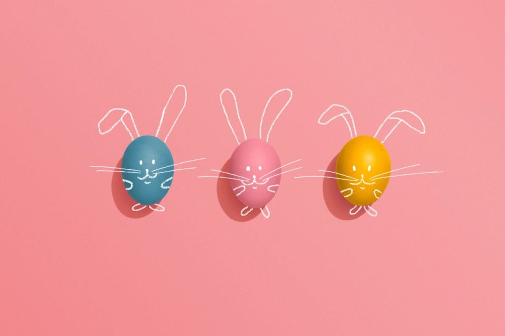 three colourful easter eggs laid out on pink paper with faces and whiskers drawn on them, and bunny ears in white on the paper