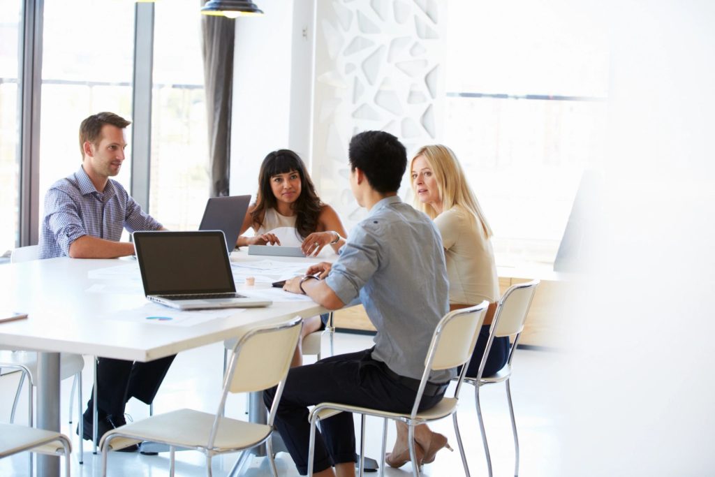 Four people sitting around a modern glass table with white leather chairs, laptops open, talking