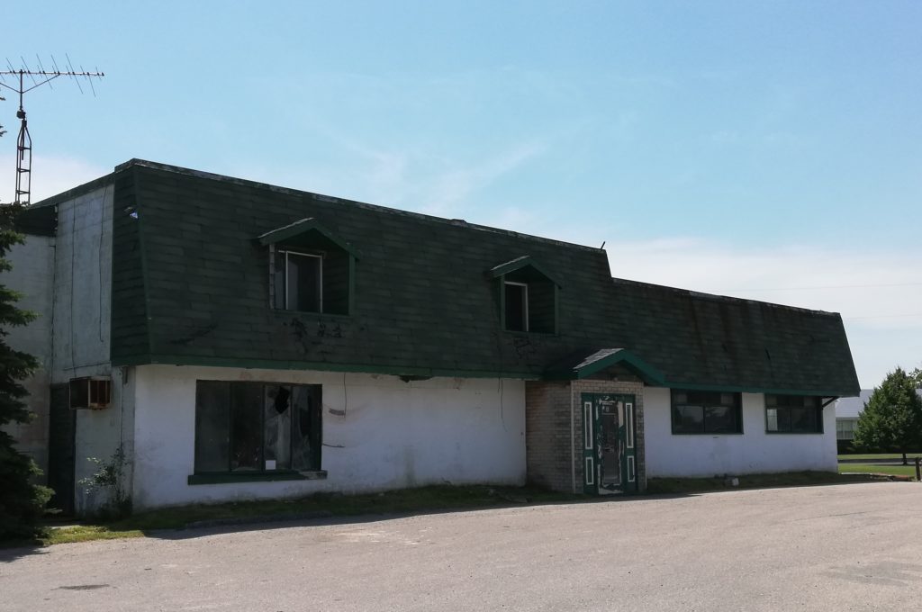 ugly old white building with a black, overhanging roof, with smashed-in windows and chipping paint