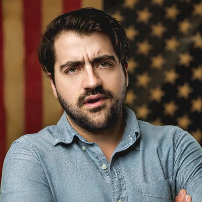 closeup of Trae Crowder from his Twitter profile - dark-haired man with a trimmed moustache and beard wearing a light denim shirt