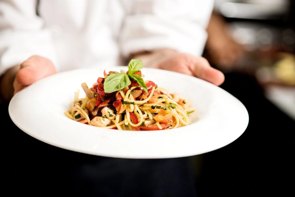 A waiter holds a white bowl full of spaghetti with red sauce and a bay leaf on top