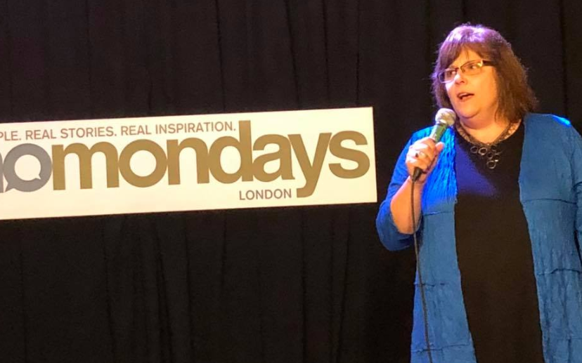 Me wearing a black dress with a bright blue duster, talking on the mic, beside a sign that reads momondays London - Real People. Real Stories. Real Inspiration.