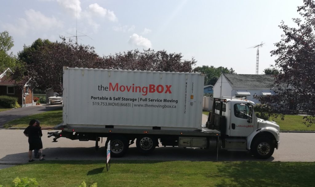 Full shot of the Moving Box seacan on a flat bed truck. Derek and the driver are discussing where it will be placed. It's white with orange and black lettering and 20 feet long.