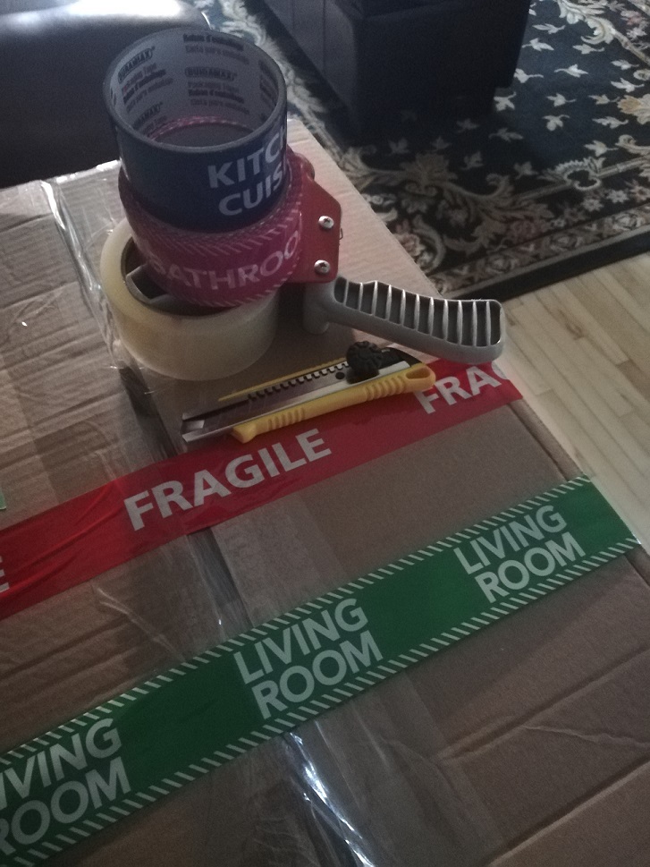 Rolls of tape reading KITCHEN and BATHROOM on top of a tape gun with packing tape. A box cutter. All on top of a packed box with tape across it - red tape reading FRAGILE and green tape reading LIVING ROOM.