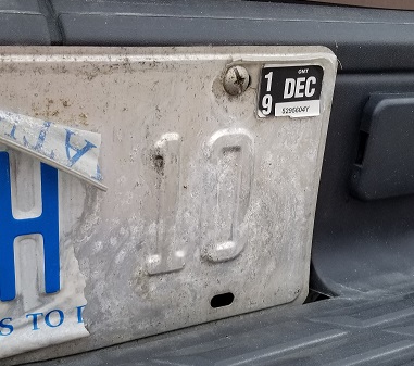 Close-up of license plate shows the coloured cover has peeled away from the 10 and it's hanging over an H, soon to come right off. 