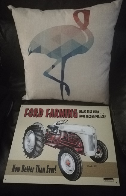 A canvas throw pillow with a pink and blue flamingo on it. A metal sign with a drawing of a Ford Ferguson tractor that reads Ford Farming means less work ...more income per acre.