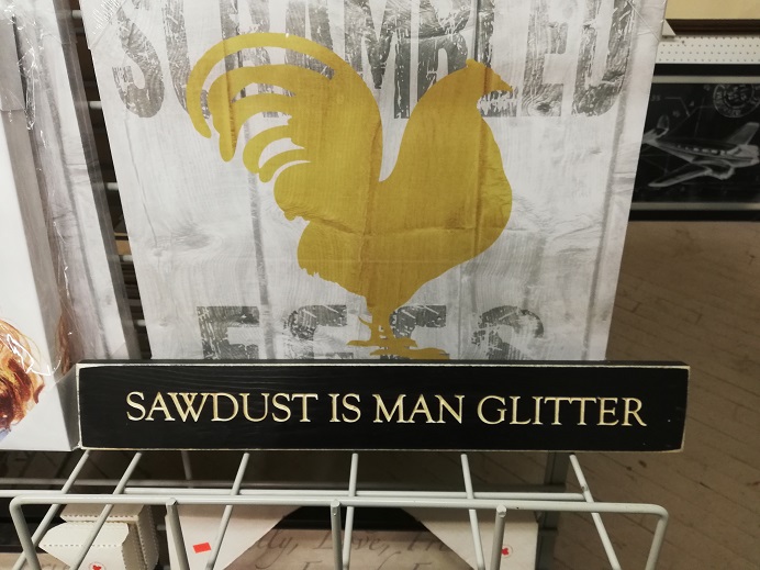 A sign with a painted chicken on it reads, Scrambled Eggs. In front of it, a long, short black sign that reads, in white, Sawdust is Man Glitter. 