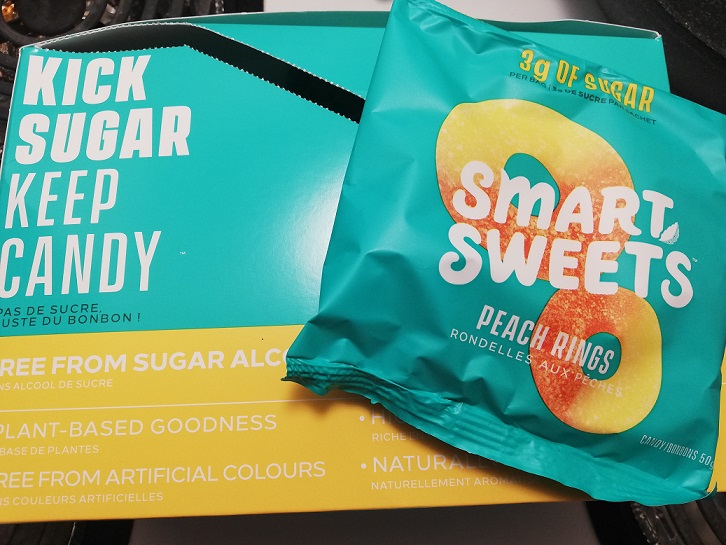 Blue and yellow box with white writing says Kick Sugar Keep Candy. On top is a package of Smart Sweets Peach Rings. 