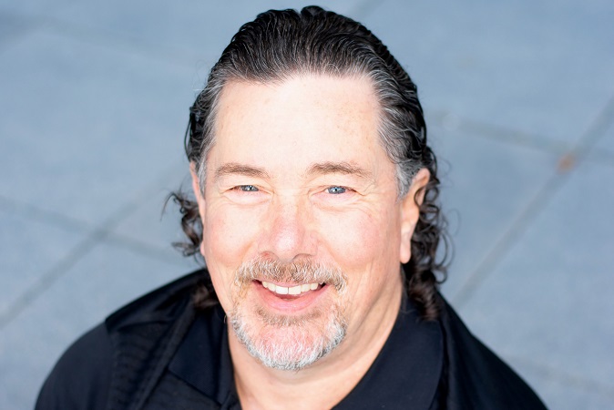 Derek in a photo taken from overhead. He's smiling, wearing a black golf shirt, and it's head and shoulders with a brick background.