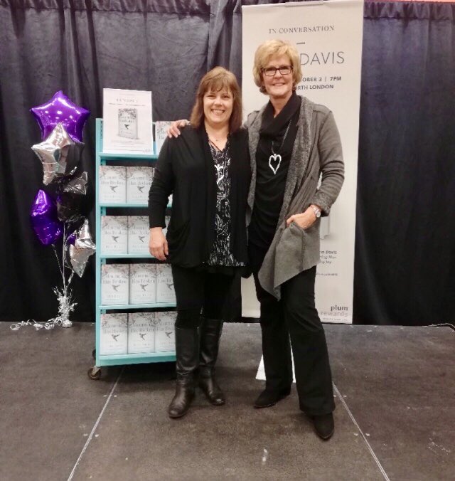 Erin and I arm in arm on the Indigo stage, in front of a shelf of her books and a large poster with the book cover on it. 