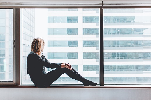 woman on a windowsill of a large room in a highrise, looking out the window