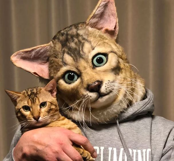 A man wears a cat mask or hood that looks just like the cat he's holding. 