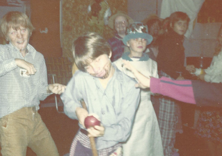 Kids dancing and having fun in costume. One in the foreground is eating an apple he just bobbed for. 