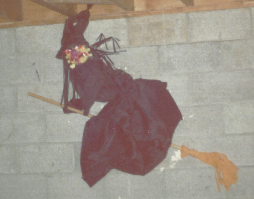 Large witch attached to the basement concrete block wall. It's wearing a black dress and flying on an actual broom affixed to the wall. 