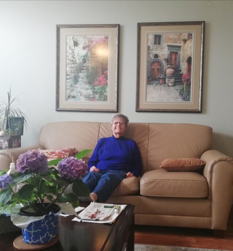 My Mom looking very tiny in a purply top and black pants sitting on a tan leather sofa below tow large prints of Italy