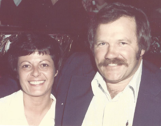 Mom and Dad smiling happily into the camera. Both around 40 years old. Mom is shorter, with short black hair. Dad is wearing a dinner jacket and has a moustache.