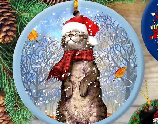 Smiling cat wearing a Santa hat and red plaid scarf in a snowstorm, on a round, blue ornament. 