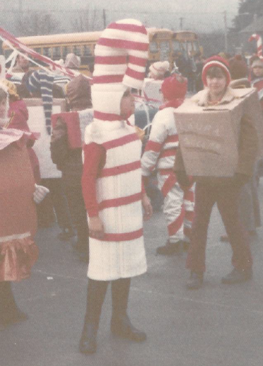 12-year-old me in a side view, wearing a white foam shell dress and a hat with red stripes. The hat curved down to my forehead, like a candy cane. 