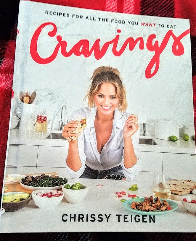 The cover of Chrissy Teigen's recipe book, Cravings, features her smiling behind a counter filled with plates of different foods.