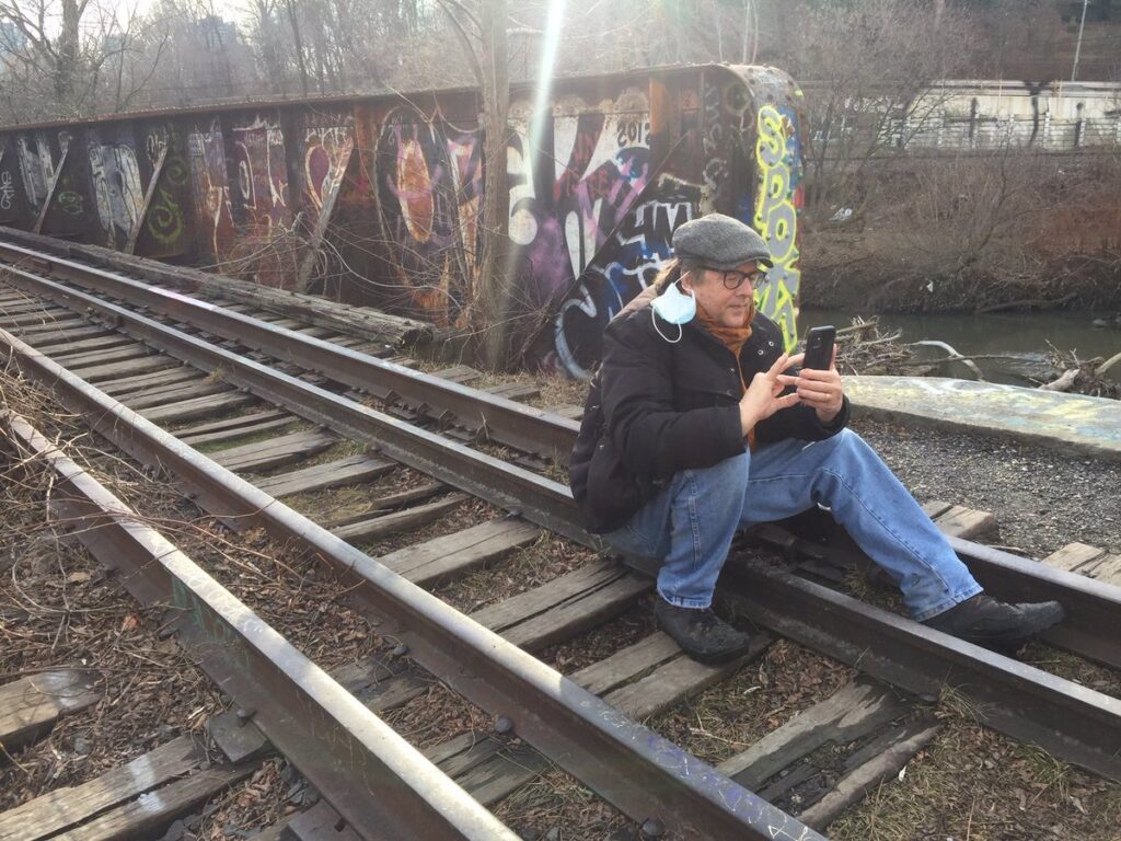 Our friend Louis with his mask hanging from one ear, sitting on train tracks looking at his phone with a wall of graffiti behind him. 