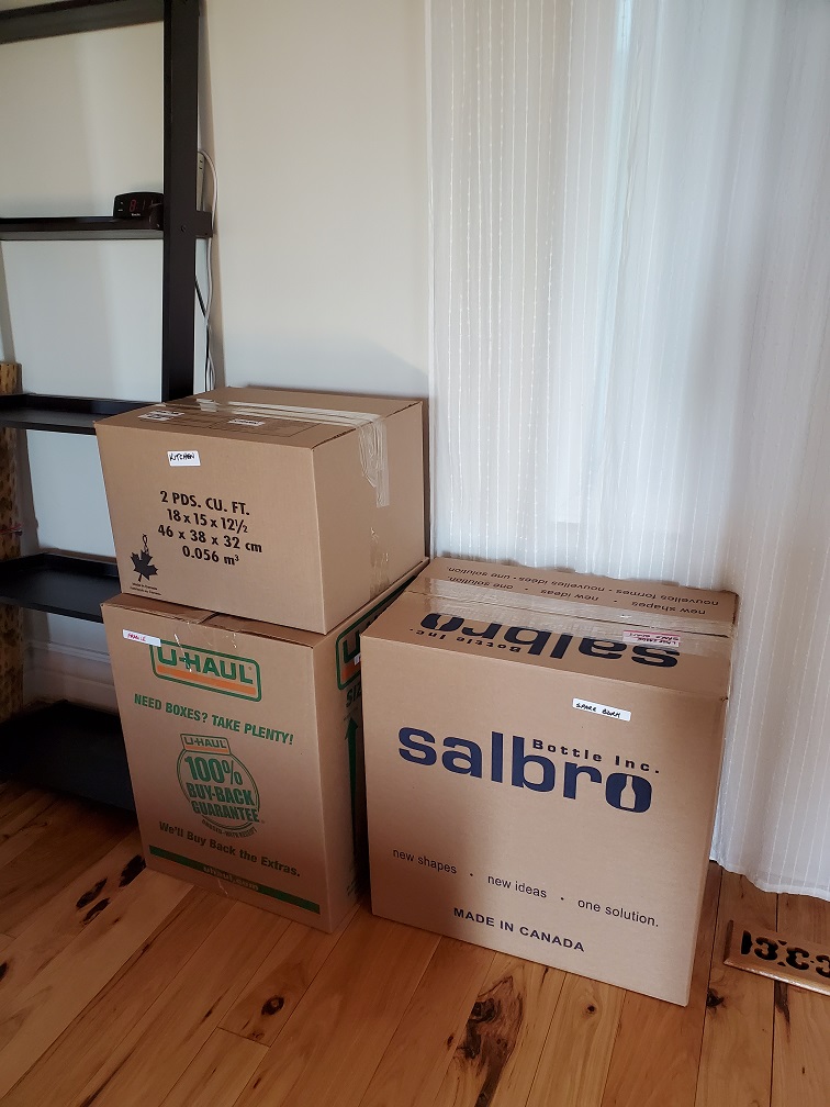 Three large, packed and sealed boxes stacked beside an empty, tall bookshelf.
