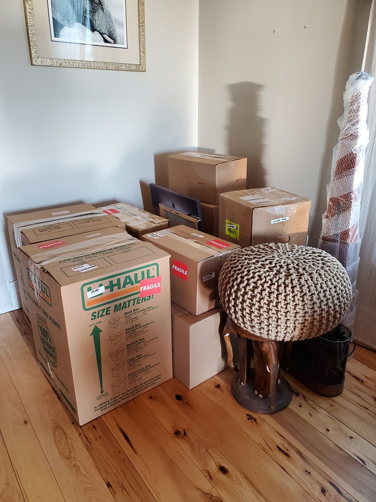A dozen or so boxes in the corner of a room. A bubble-wrapped lamp. A pouf on top of a footstool. Some pictures stacked on end. 