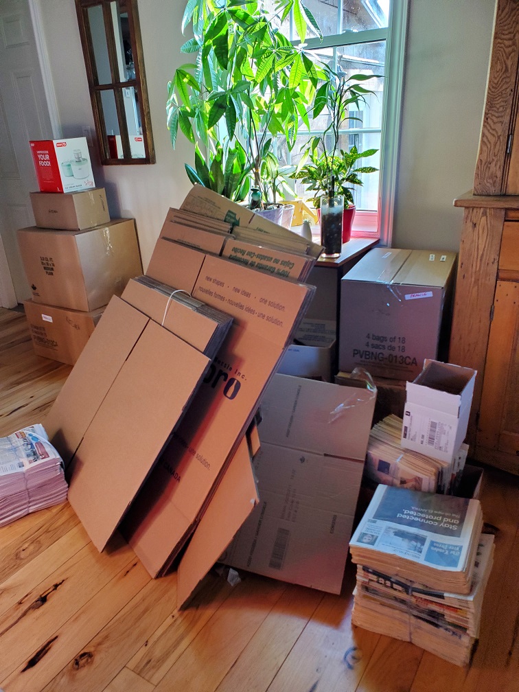 Flat boxes stacked against a table full of plants next to a window. Huge bundles of newspapers. Five packed boxes stacked on the far left.