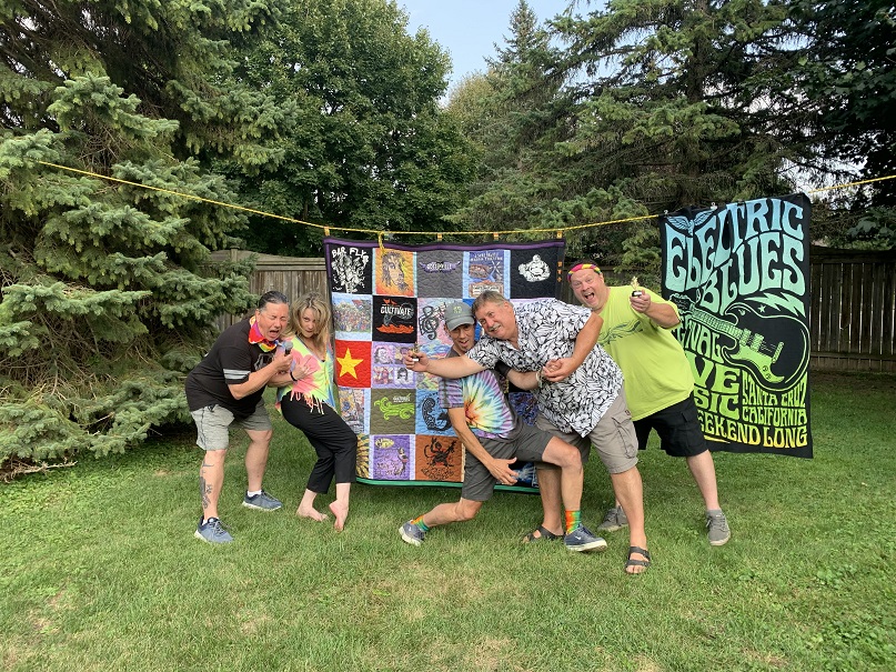 The five remaining Botten siblings all taking rock-star poses in front of a large quilt hanging on a clothes line. They're dressed in tie-dye colours and outside on grass with tall evergreens in the background.