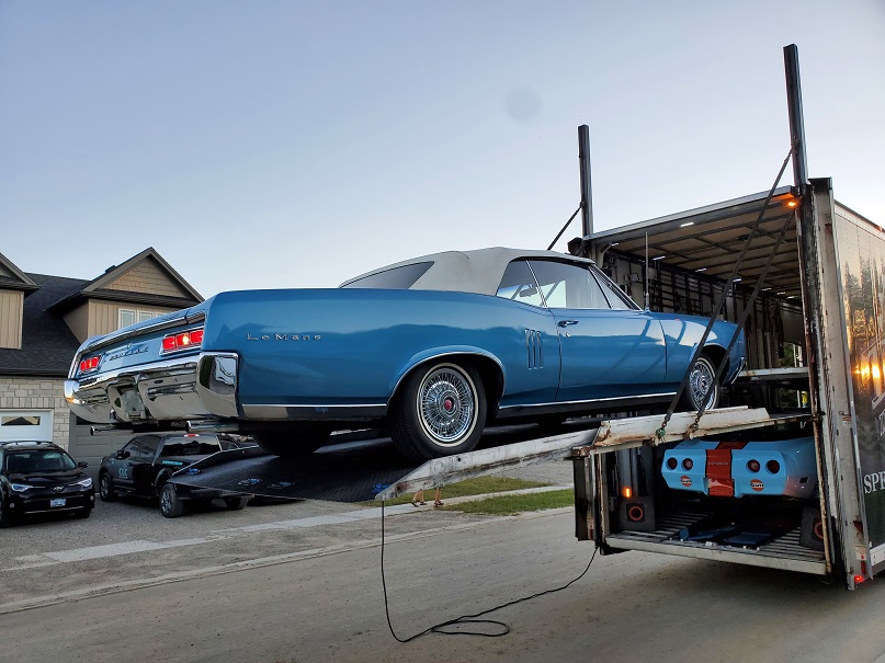 Bright blue Le Mans ragtop convertible suspended off the back of a tractor trailer on a metal tray. 