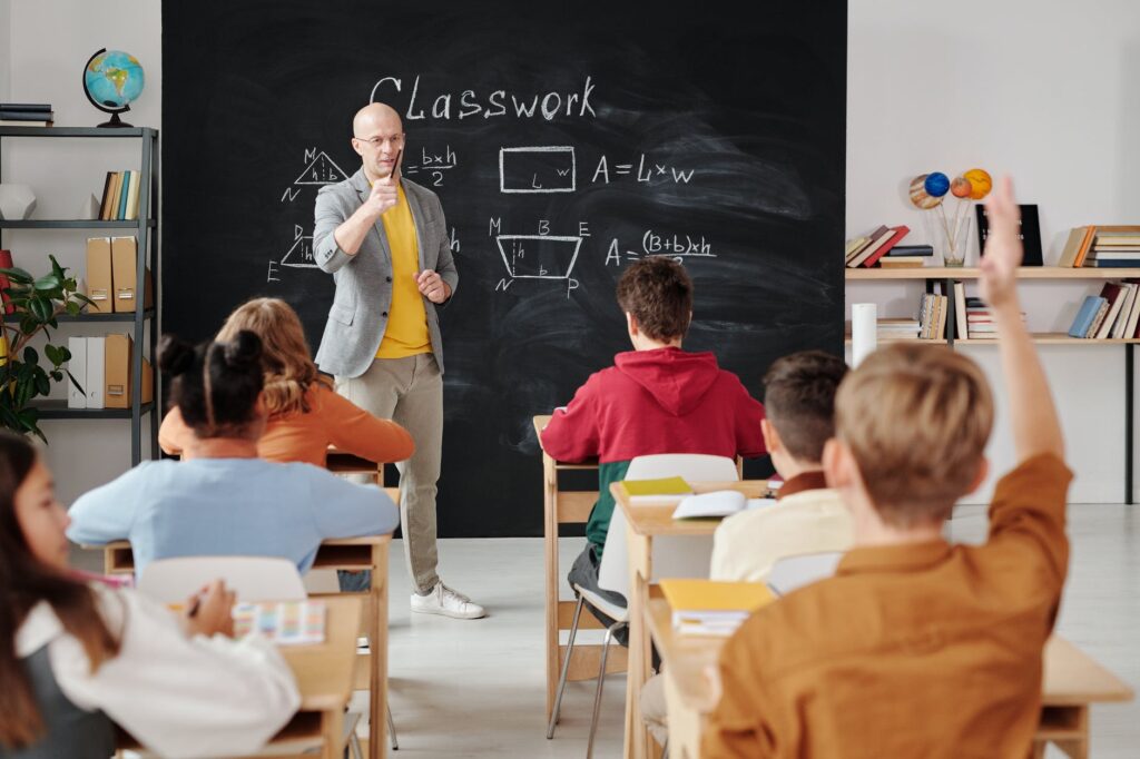 Teacher in front of a blackboard teaching a lesson, gesturing toward students sitting at their desks.
