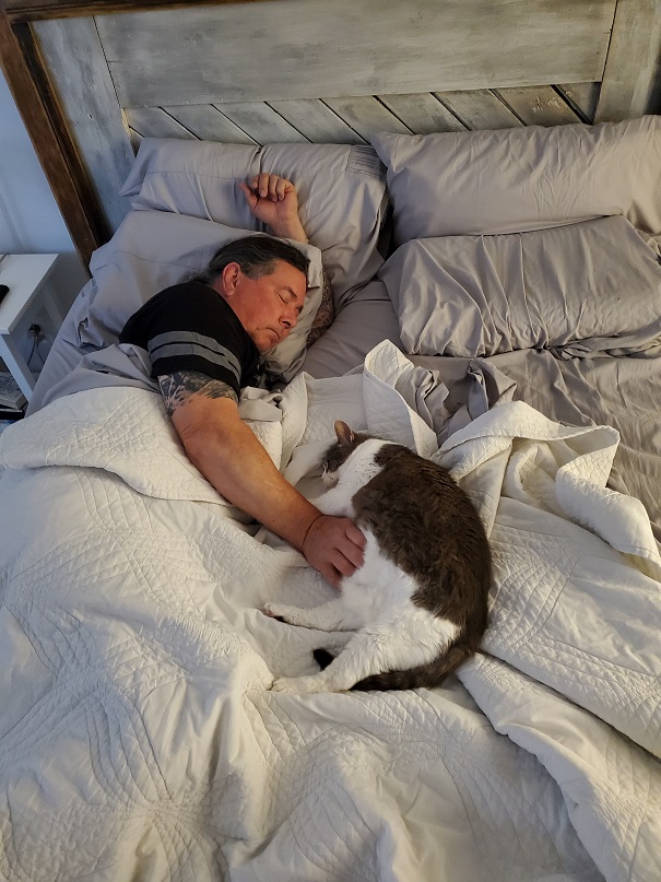 Derek and Sugar asleep on a rumpled bed, facing each other. He's on his side with his hand on her belly and she's also fast asleep on her side. 