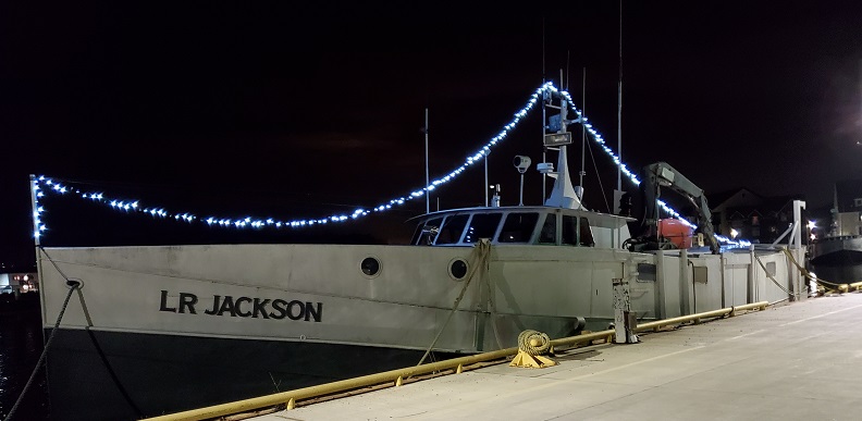 The LR Jackson, a big fishing boat, docked at the harbor with clear lights strung from one end to the other, attached to the mast. 