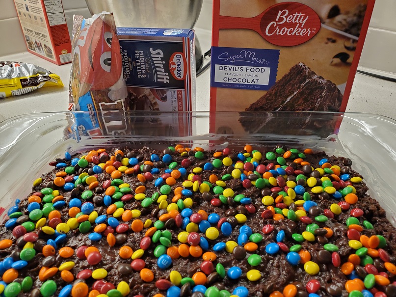 Glass rectangular pan with chocolate cake covered in M and M minis. Behind it are a box of Duncan Hines cake mix, a box of pudding mix and the M and M minis bag. 