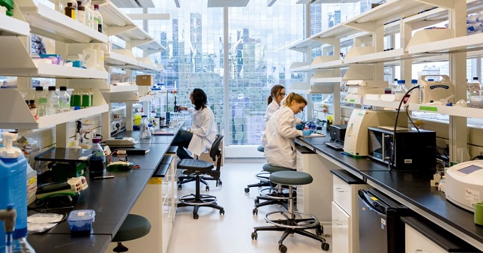 long view of a research lab with all sorts of lab stuff on a long counter and on high shelved with three scientists at work