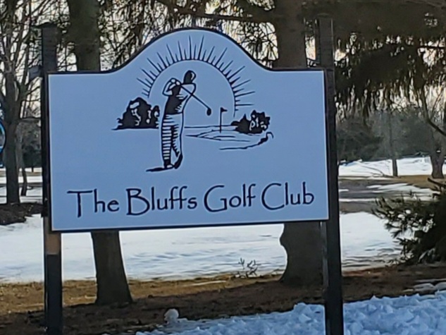 Sign for The Bluffs Golf Club. White signs with black lettering and a drawing of a man hitting a ball with bright sunshine in the distance. 