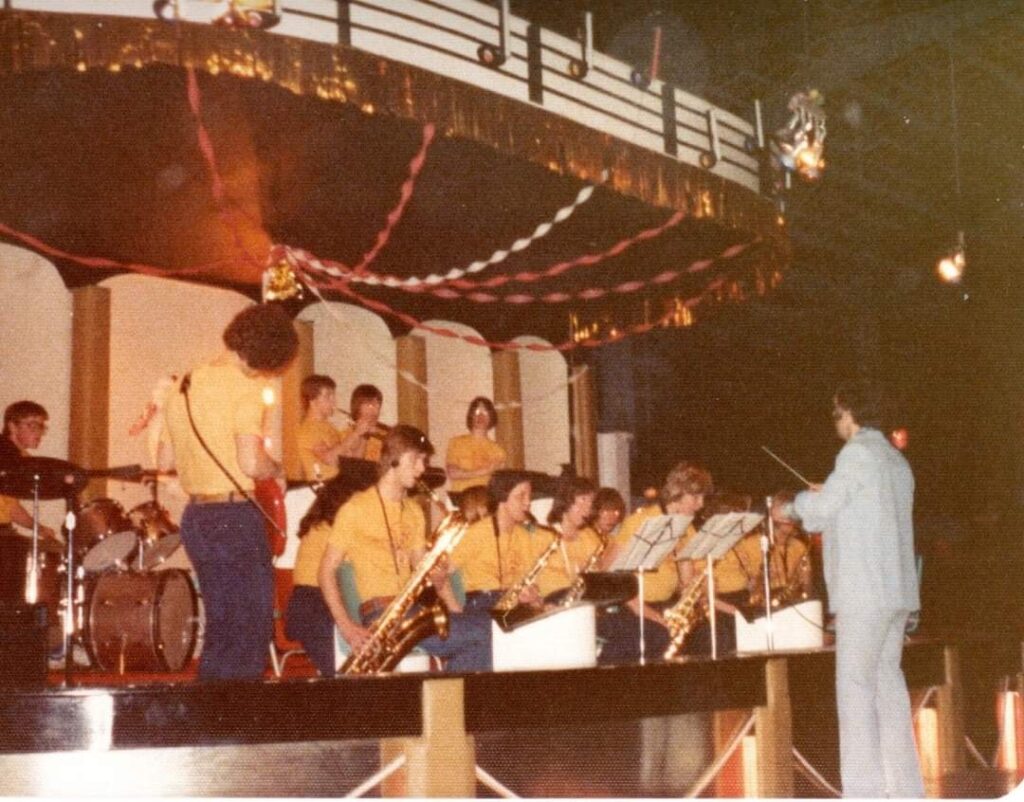 A teenage band all wearing gold T-shirts and jeans, playing in an orchestra. The conductor is in a light blue suit. 