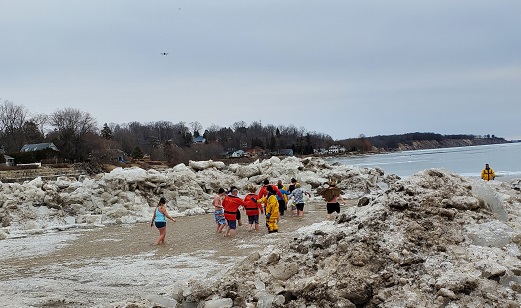 Five people in bathing suits enter the icy water with banks of snow on either side. Firefighters in thermal suits watch over them. 