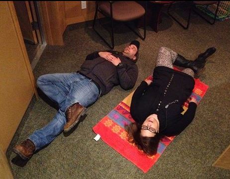 Blue-jean wearing Blair on his back on the carpet, and me beside him on a beach towel. We both have one leg crossed over the other as I show hiim stretches. 