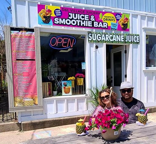 A couple sits at a picnic table in front of D & L which is a white building with colorful banners listing their menu out front. 