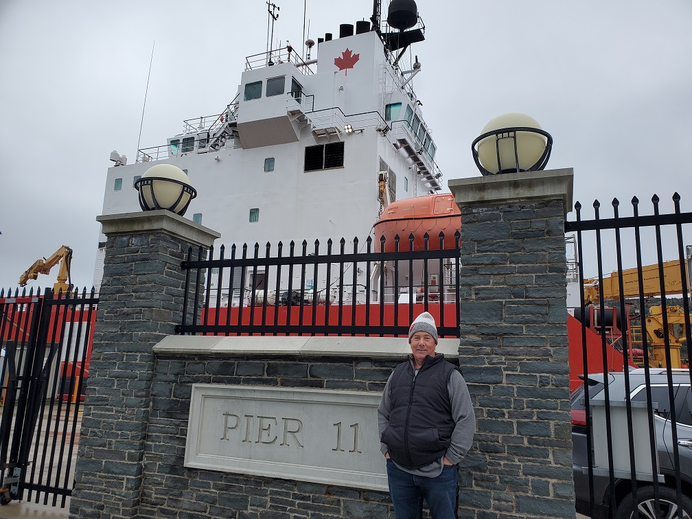 Derek in front of a brick wall amid an iron fence that reads Pier 11 in St. John's Newfoundland. Behind him is a huge Canadian government ship.