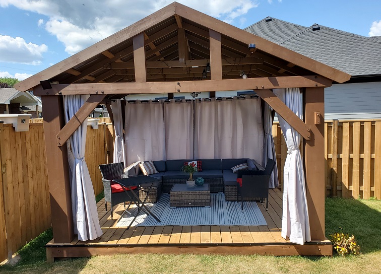Wooden gazebo with a peaked roof and inside, a grey sectional, blue-gre rug, and curtains all around, tied back on the front and sides.