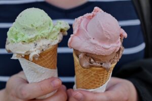 two ice cream cones held to the camera by an unseen person. One has mint and vanilla. The other is strawberry on top of chocolate.