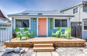 Small blue cottage in Port Stanley with a wooden deck out front and green chairs on it. The cottage has an orange door.