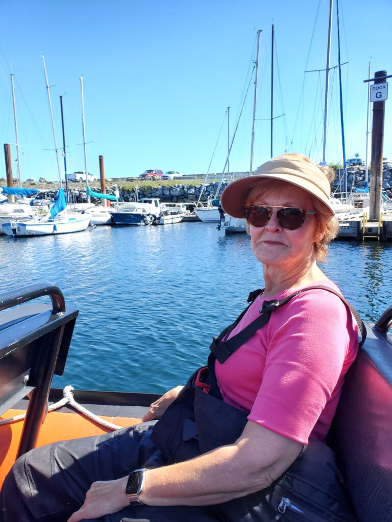 Erin sitting in our whale-watching boat, wearing what looks like fishing waders and suspenders over a pink T shirt.