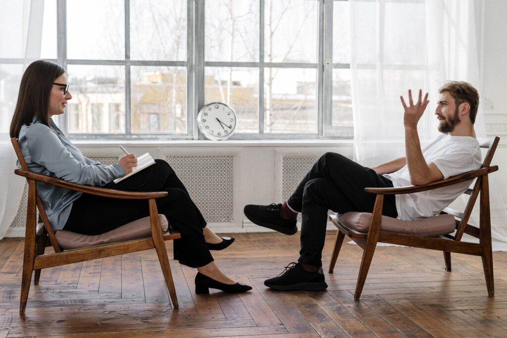 Person in Black Pants and Black Shoes Sitting on Brown Wooden Chair talking to female therapist opposite him