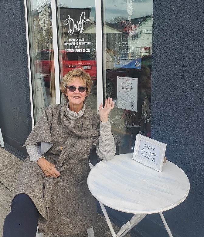 Erin sitting at a little bistro set outside a shop identified as Drift. On the bistro table sits a sign that reads, Drift Husband Parking. 