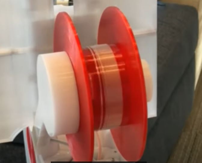 Red spool with clear filament which is a rounded strip of plastic from a water bottle. 