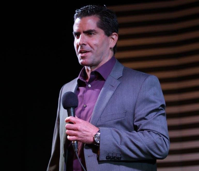 A man in his 40s holding a microphone, wearing a purple shirt and blue jacket.
