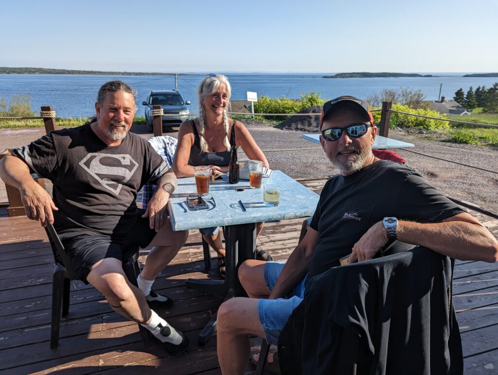 Derek, Monique and Tam sitting on a deck with beverages on the table and the ocean in the background. 